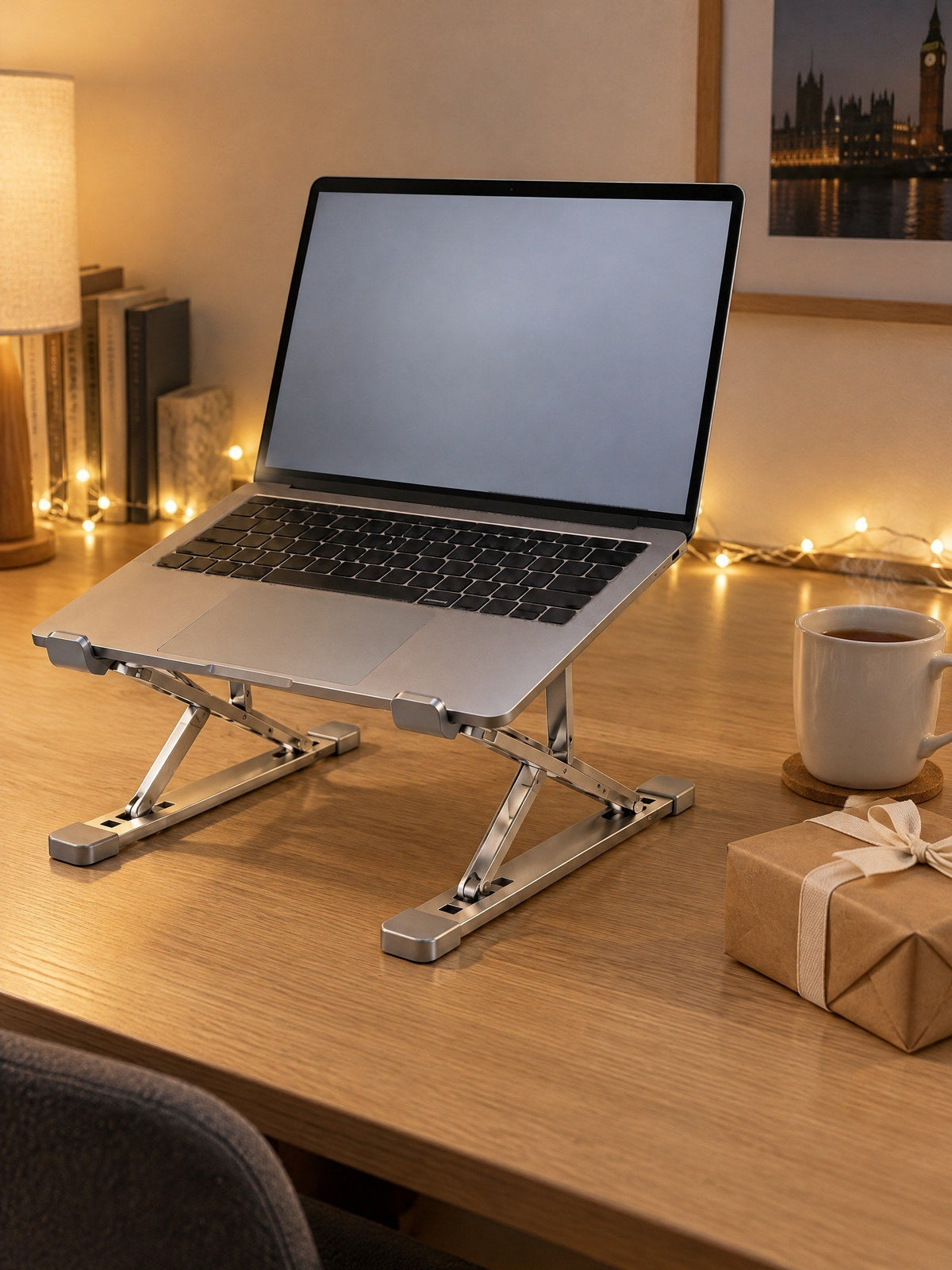 Silver folding laptop stand, Tall model, holding an open laptop on an oak desk with fairy lights, a mug and a wrapped present