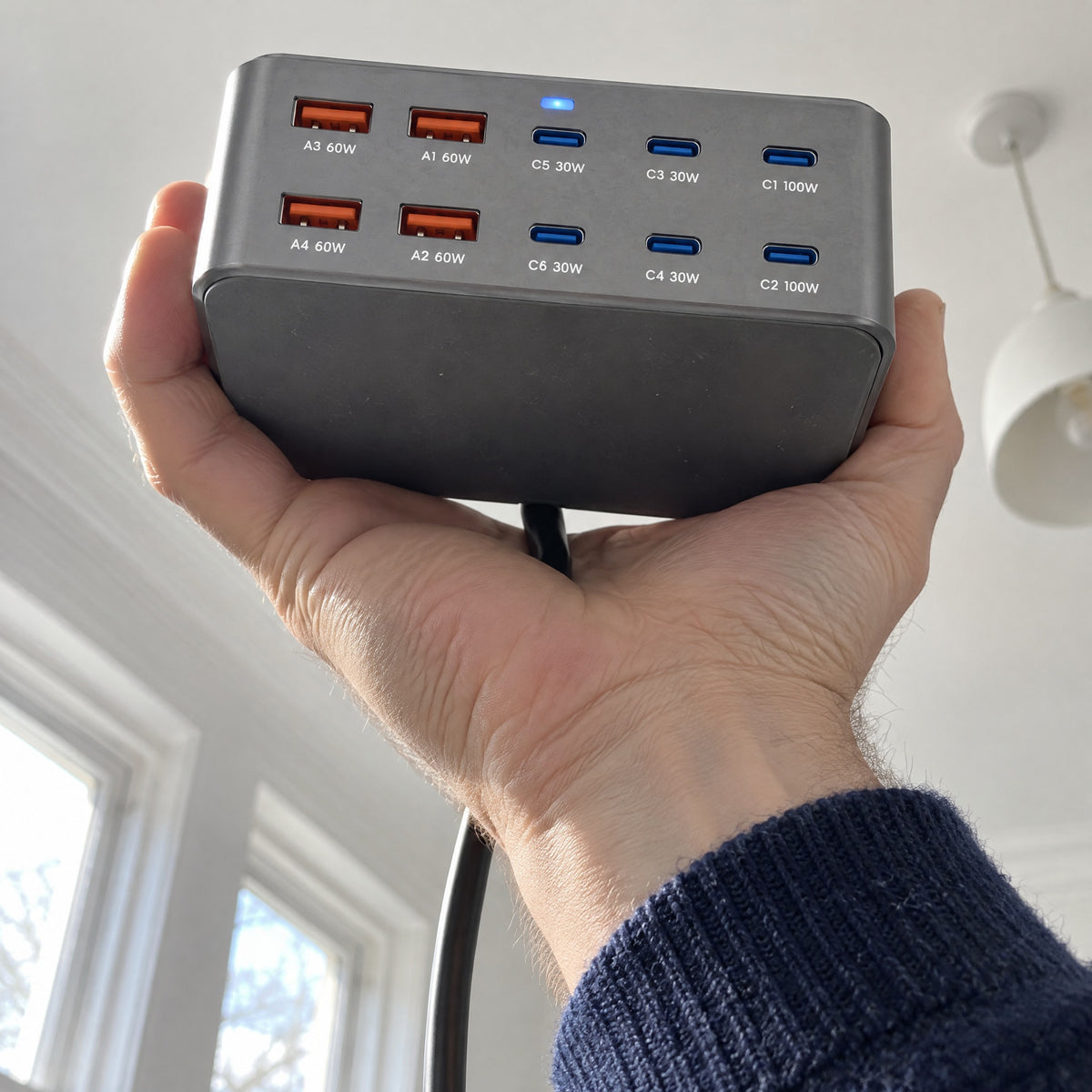 Wharf charger held overhead and seen from below, its ten labelled USB-A and USB-C ports facing the camera, black mains cable hanging behind
