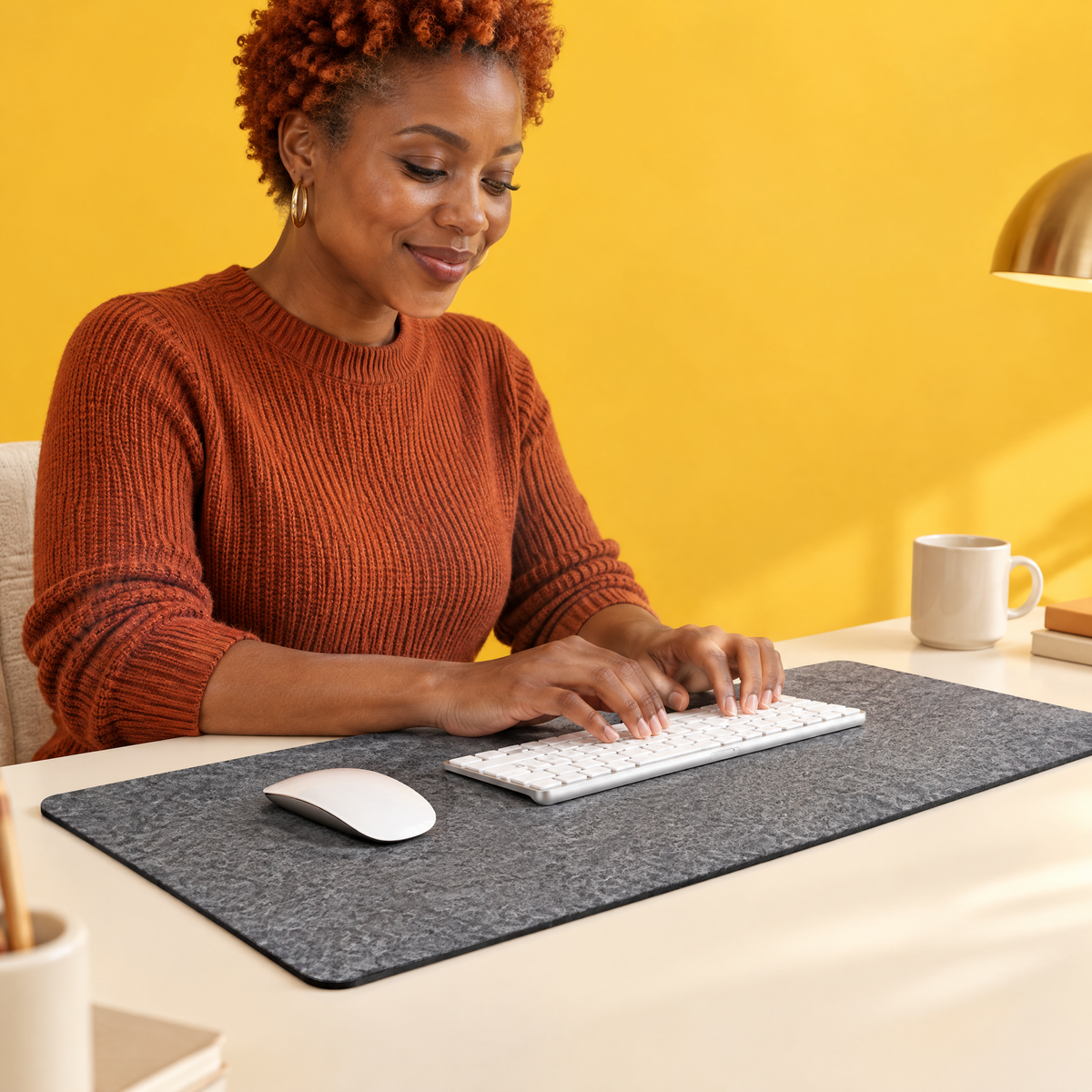Woman in a rust-red jumper typing on a white keyboard on a grey felt desk mat, with a white mouse beside it and a yellow wall behind