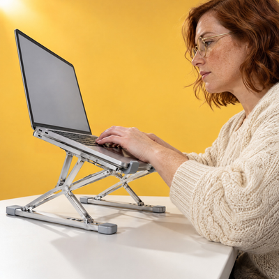 Woman typing on a laptop raised on the Tall folding aluminium stand in silver, seen from desk level