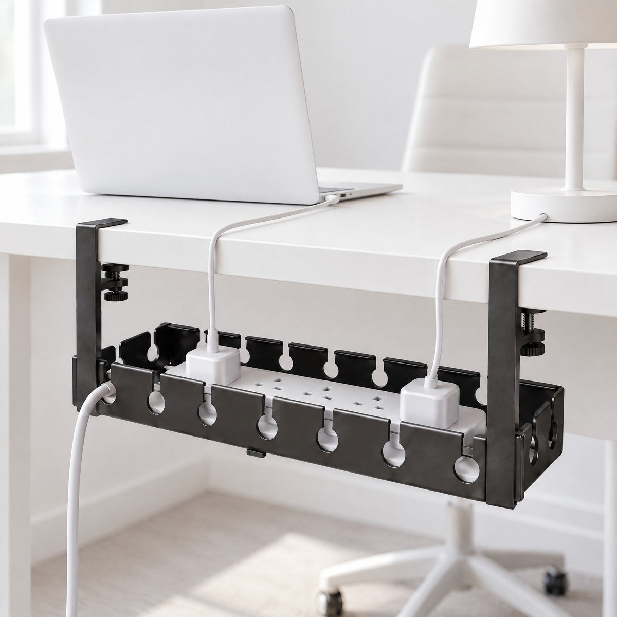 Black Hideaway cable tray clamped under the back edge of a white desk, holding an extension lead and two chargers