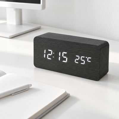 Black Grain wood-effect LED clock showing the time and temperature on a white desk beside an open notebook