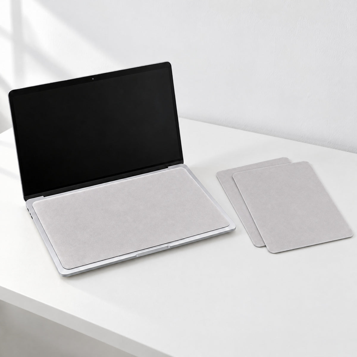 Light grey microfibre keyboard cover cloth lying over a silver laptop keyboard, two spare cloths stacked beside it on a white desk