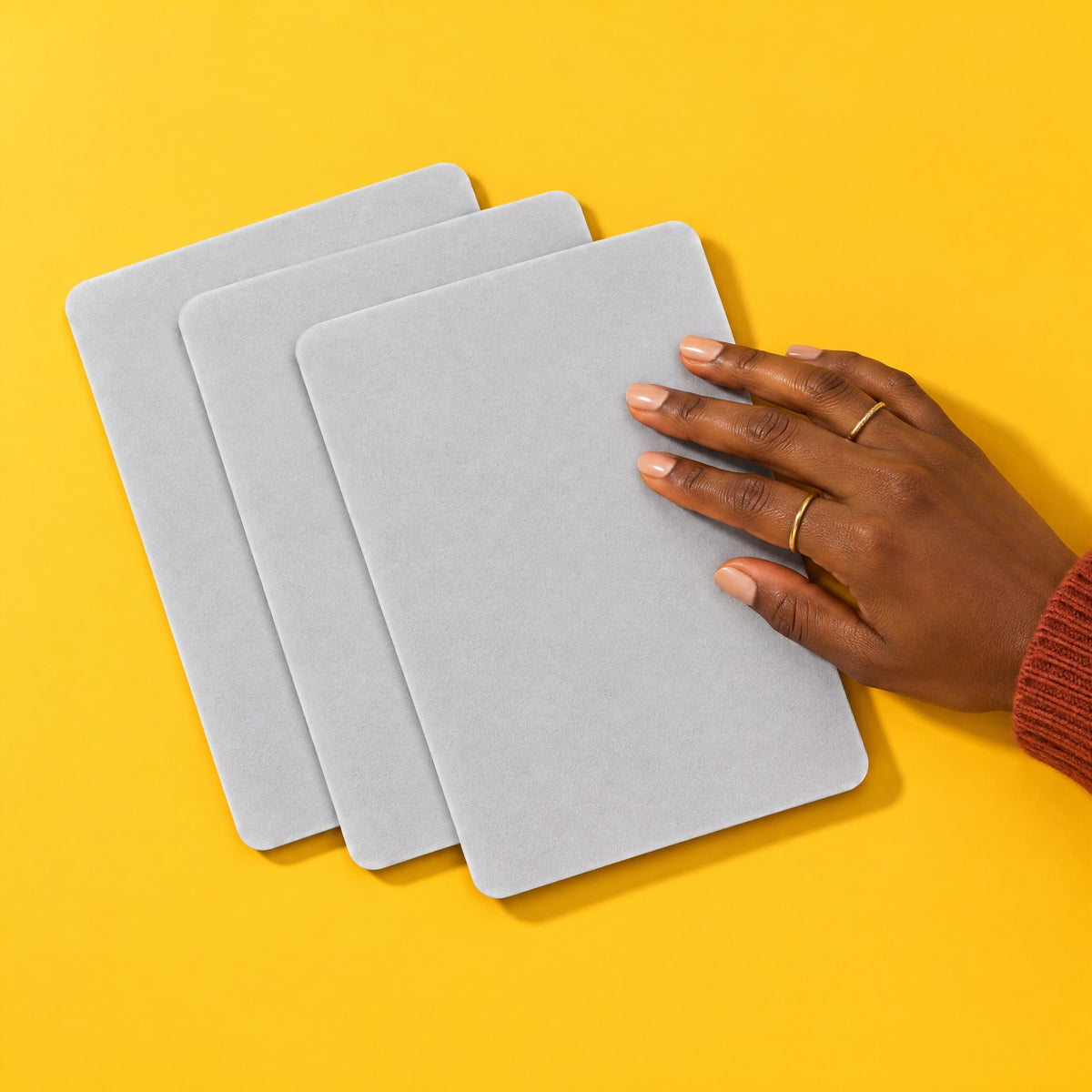 Pack of three light grey microfibre keyboard cover cloths fanned out on a yellow backdrop, a hand sliding the top one into place