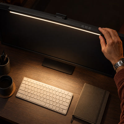 Hand tilting the Crest light bar on top of a monitor, its warm light falling on the keyboard below