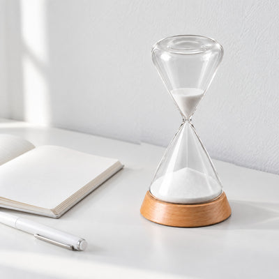 Ebb glass hourglass with white sand in a beech base on a white desk beside an open notebook