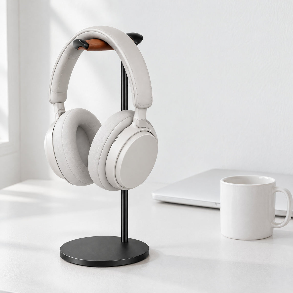 Black aluminium headphone stand with a tan leather rest holding white over-ear headphones on a white desk
