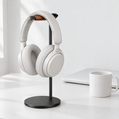 Black aluminium headphone stand with a tan leather rest holding white over-ear headphones on a white desk