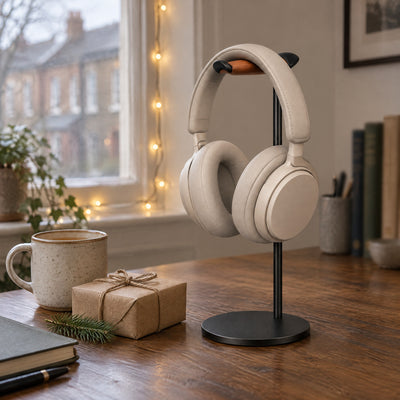 Black headphone stand with cream headphones on a wooden desk beside a kraft-paper wrapped gift and a pine sprig