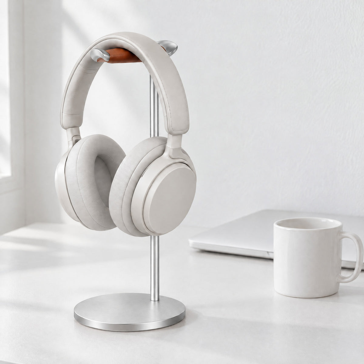 Silver aluminium headphone stand with a tan leather rest holding white over-ear headphones on a white desk