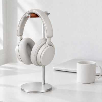 Silver aluminium headphone stand with a tan leather rest holding white over-ear headphones on a white desk