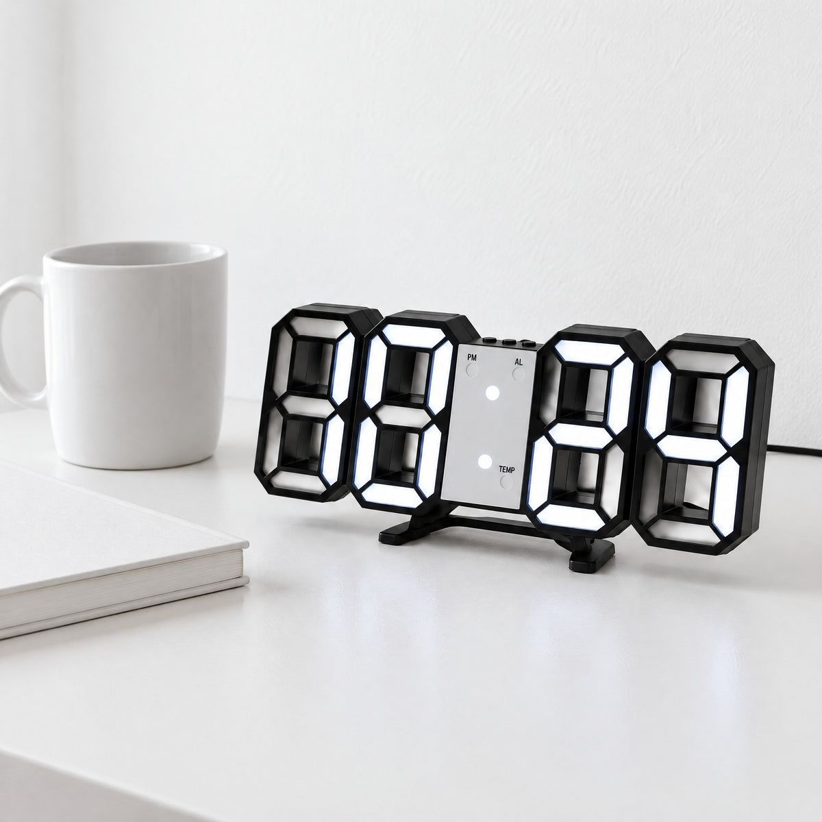 Black 3D LED digital clock with white light standing on its fold-out stand on a white desk beside a mug and notebook