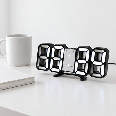 Black 3D LED digital clock with white light standing on its fold-out stand on a white desk beside a mug and notebook