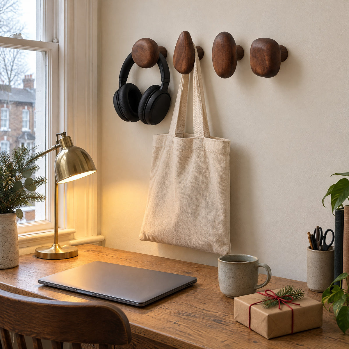 Walnut pebble wall hooks holding headphones and a canvas tote beside a home-office desk with a small wrapped gift