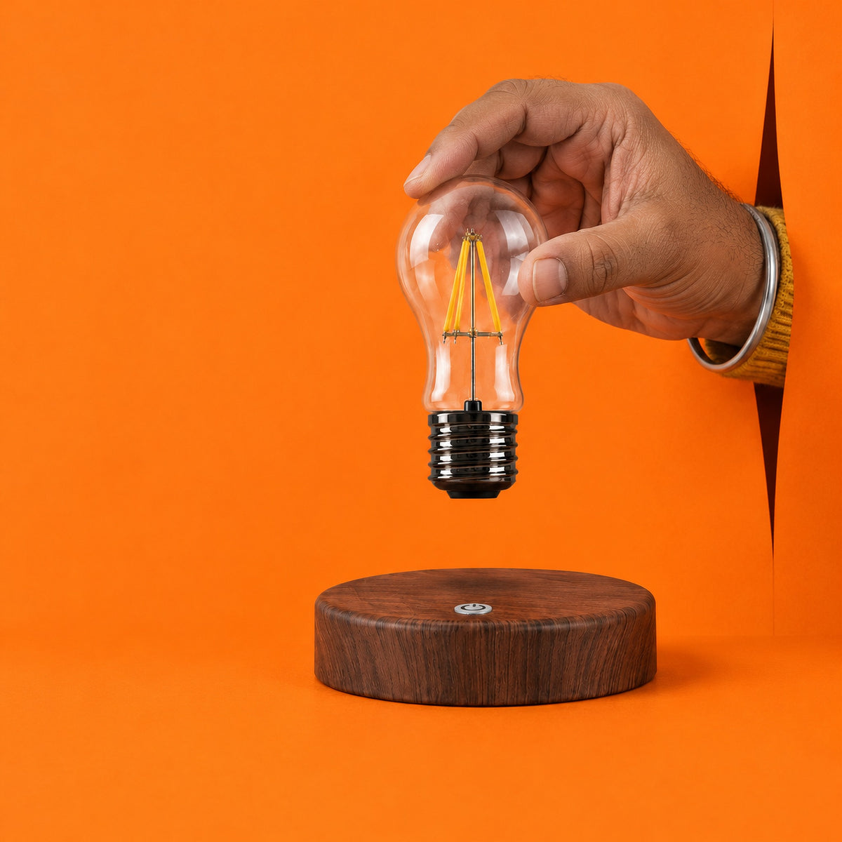 A hand lifting the clear glass bulb of the Drift lamp off its round base, against an orange backdrop