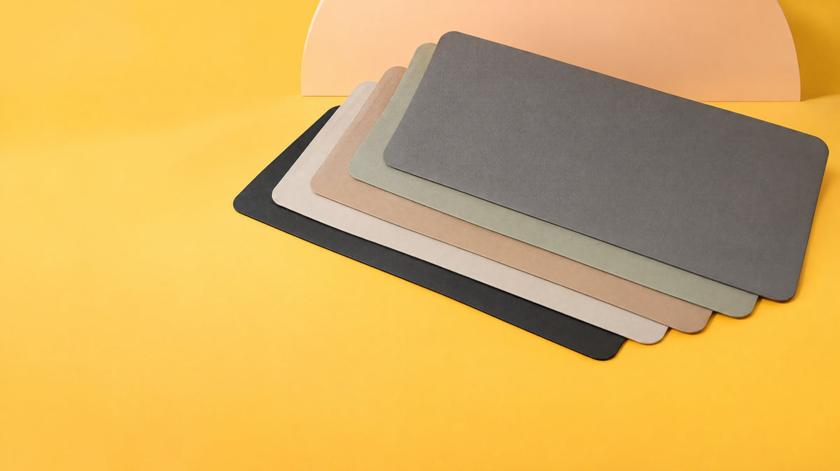 Five faux leather desk mats fanned out in black, eggshell, latte, sage and dark grey on a sun-yellow backdrop
