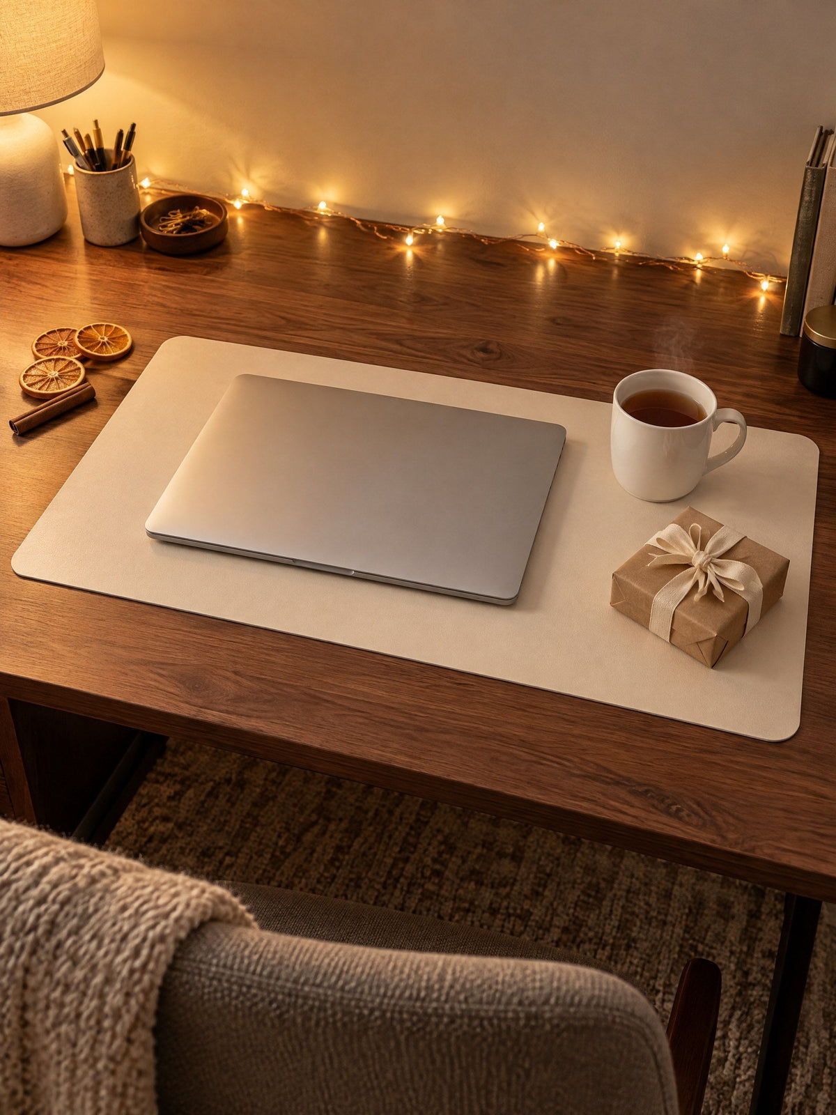 Eggshell desk mat on a walnut desk in the evening, with a closed laptop, a mug of tea, a small wrapped present and fairy lights