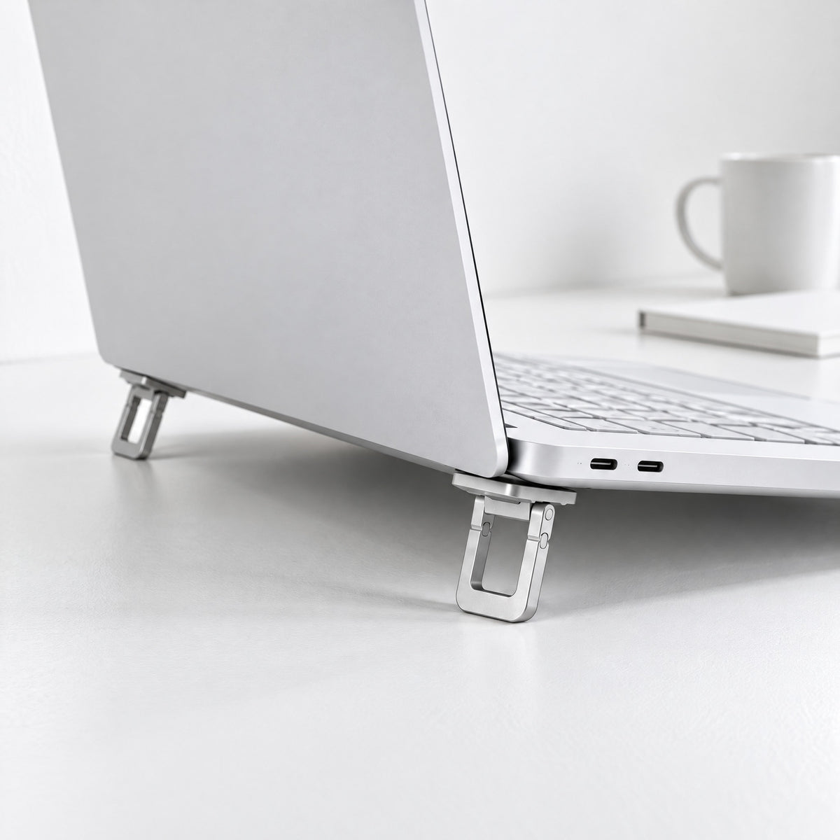 Stilt folding laptop feet in silver, open under the back corners of a white laptop on a white desk