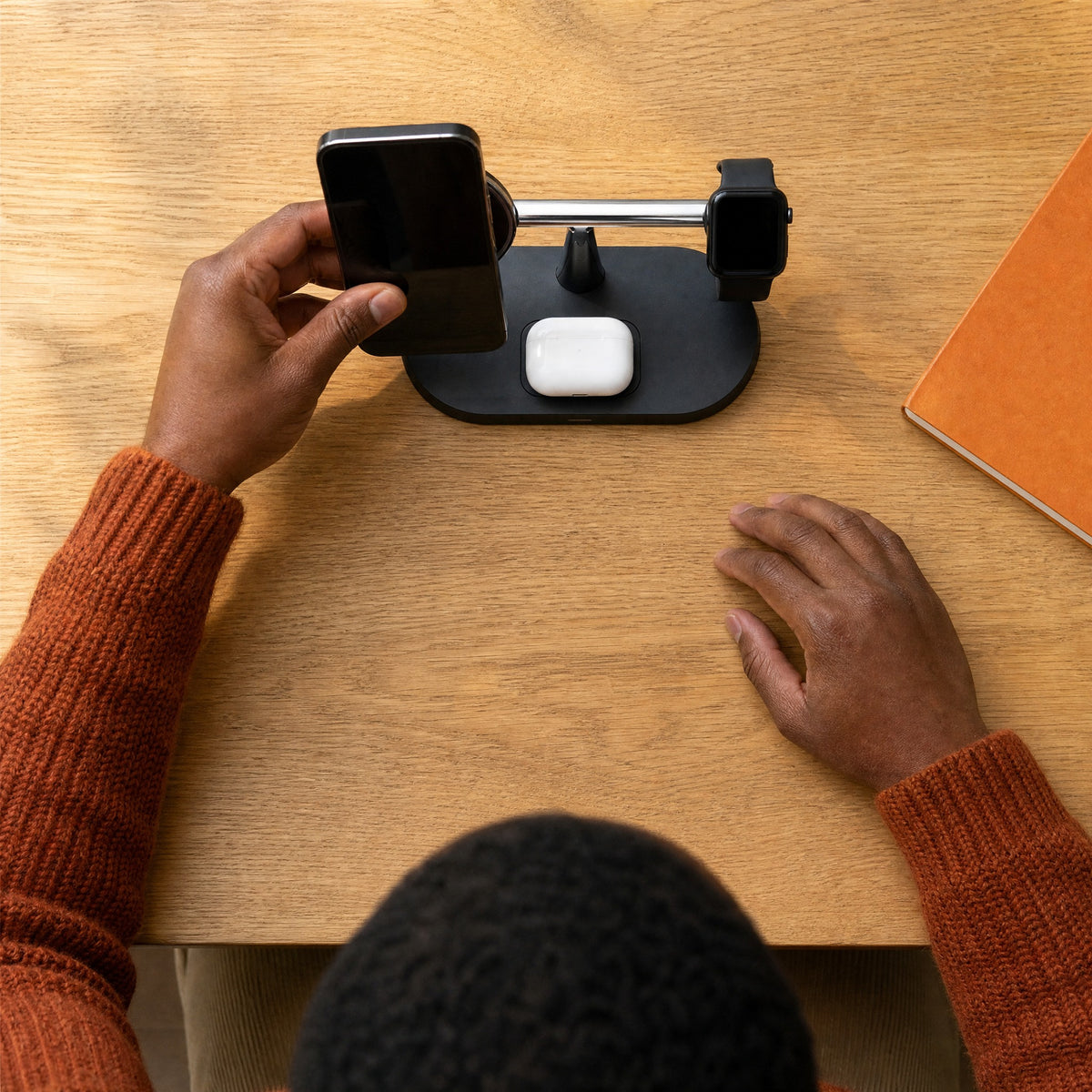 Man in an orange jumper placing his phone on the black Trio charging stand, seen from above, with a watch and an earbuds case already on it