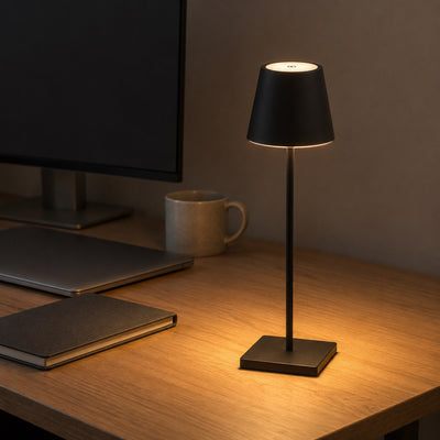 Black Vesper cordless table lamp casting warm light on an oak home-office desk beside a monitor