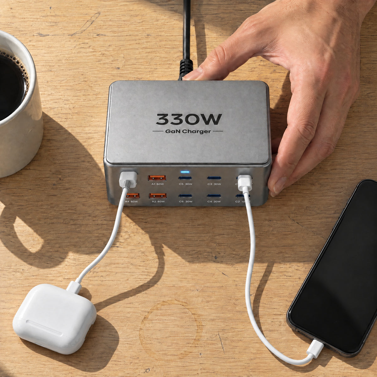 Top view of a hand setting the grey Wharf 330W charger on an oak desk, charging earbuds and a phone, black mains cable at the back
